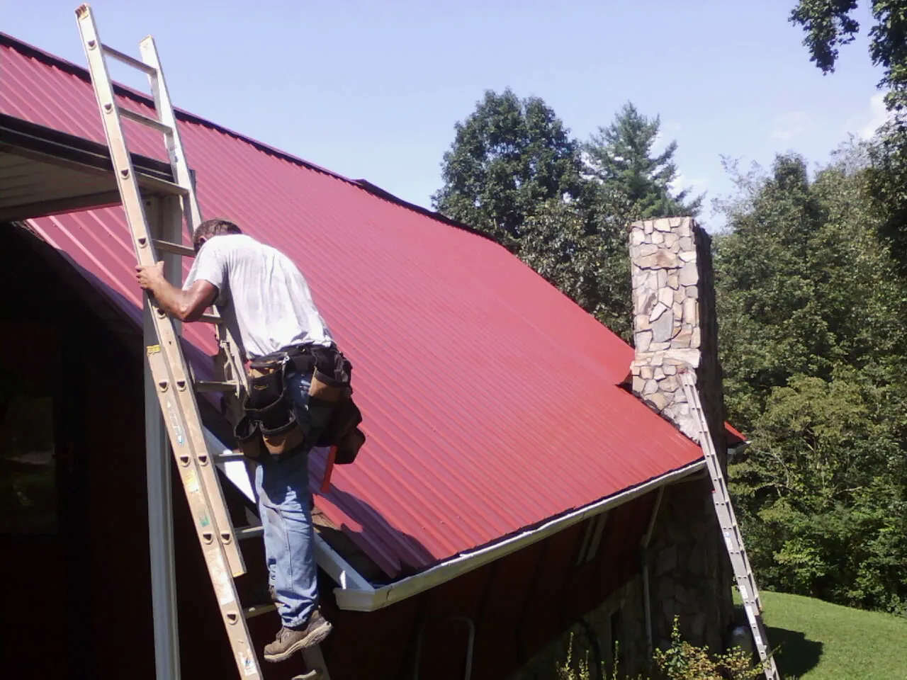 Roofer on a ladder beside a red metal roof and stone chimney — Soffit & Fascia Repair by Roofing Brothers in Checotah